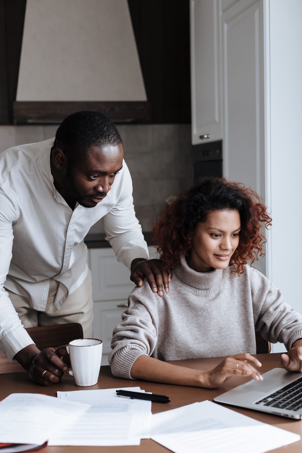 A man leaning over his partner's shoulder with a hand resting on it while she works at a laptop at home.